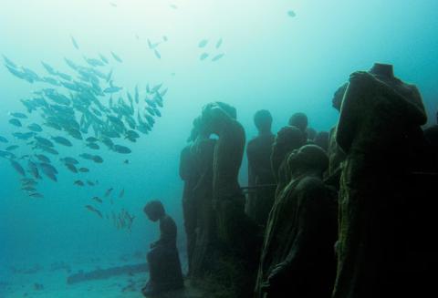 Underwater scene with weathered human statues and a school of silver fish swimming nearby.