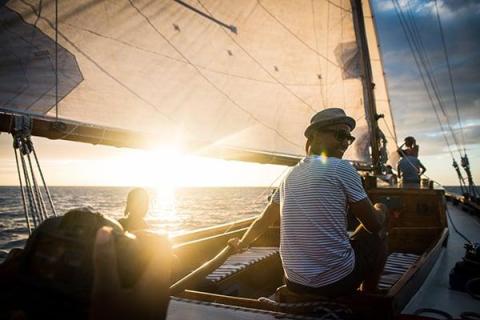 Group on sailboat at sunset with person playing piano, serene sea backdrop.