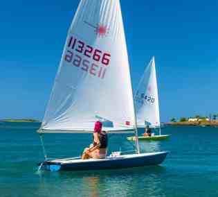 Two sailors enjoy a sunny day on calm sea with clear skies and scenic backdrop.