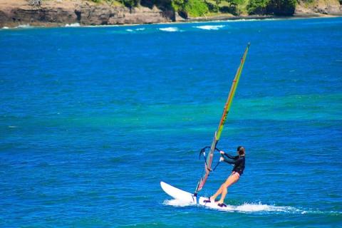 Windsurfer with colorful sail glides on blue water near rugged coast.