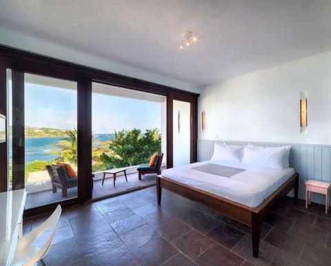 Ocean view bedroom with large bed, minimal decor, and panoramic coastal landscape.