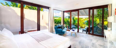 Luxurious bedroom with plush blue armchair and tropical view through floor-to-ceiling windows.