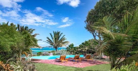Luxurious oceanfront patio with wooden sun loungers and tropical plants, facing turquoise sea.