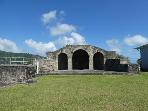 Small rustic stone pavilion with three arches, set in grassy area under clear blue sky.