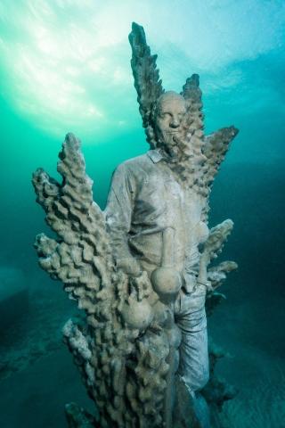 Underwater Sculpture Blending Humanity and Nature with Coral-like Structures Underwater human statue enveloped by coral-like structures, illuminated by natural light.