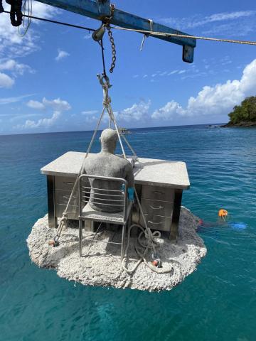 Installing Underwater Sculpture of Seated Figure with Typewriter Concrete sculpture of a person at a desk being lowered into the water by crane.