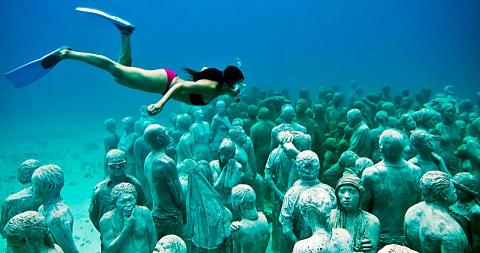 Diver explores life-sized human statues in an underwater scene with a serene, eerie atmosphere.