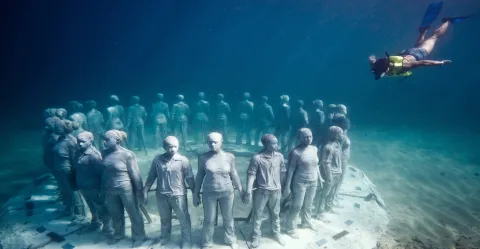 Diver Explores Serene Underwater Circle of Life-Sized Human Statues Diver swims above circular life-sized human statues on an ocean floor with marine growth.