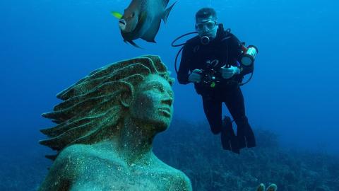 Scuba Diver Photographs Mysterious Underwater Statue amid Marine Life Scuba diver photographing a large, encrusted underwater statue with serene expression.