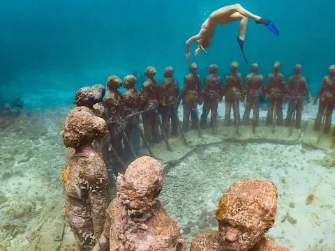 Underwater Sculptures: A Snorkeler Snorkeler swims above a circle of life-sized, algae-covered human statues underwater.