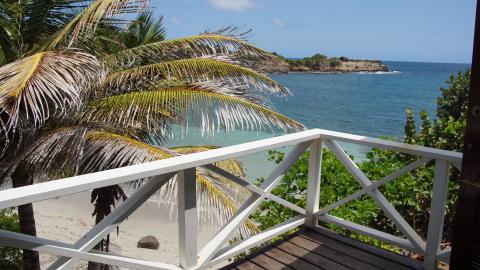 Scenic beach view from a deck with white railing, clear blue sea, and lush greenery.