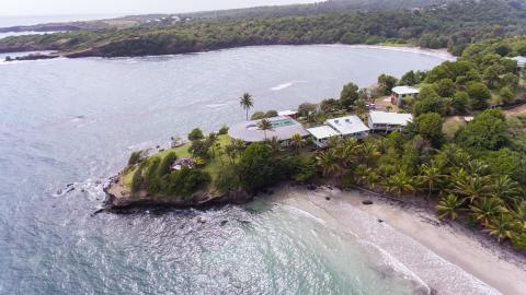 Aerial view of a secluded beach with grey sand, palm trees, and red-roof buildings.