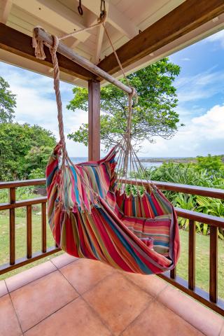 Colorful hammock on patio with ocean view, sunny sky, and lush greenery, inviting relaxation.