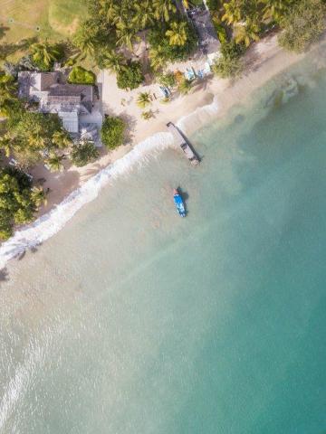 Aerial view of a sandy beach with turquoise waters, palm trees, and a small boat near dock.
