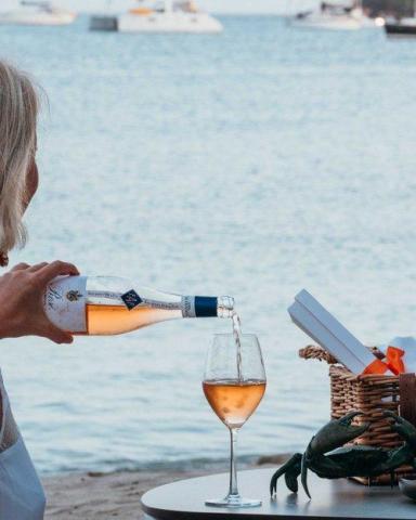 Woman pouring rose into glass by beach with boats, basket, and lobster decor.