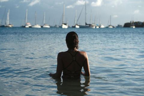 Woman in black bikini stands in ocean watching distant sailboats