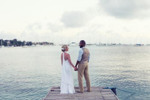 Couple in wedding attire on pier overlooking harbor with boats, soft lighting.