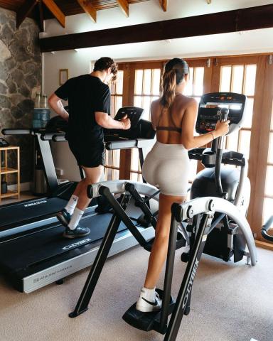 Man on treadmill and woman on elliptical in a modern-rustic home gym with Cybex equipment.
