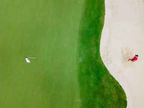 Cayman Tourism Board Precision in Play: Golfer in Red Takes a Swing from the Sand Bunker This image shows an aerial view of a golf course