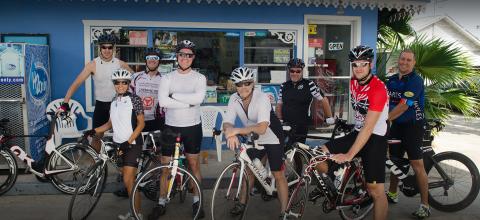 This image features a group of seven cyclists standing together beside their bicycles outside a small, cheerful blue store