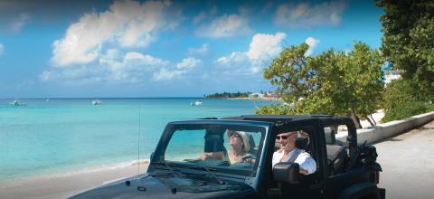 In this image, there are two adults driving along a picturesque coastal road in a convertible jeep