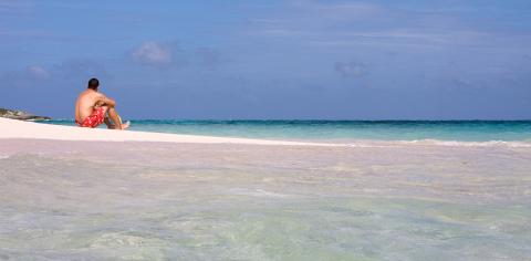 Cayman Tourism Board Solitude by the Sea This image features a tranquil beach scene with a person sitting on a pristine white sandy shore