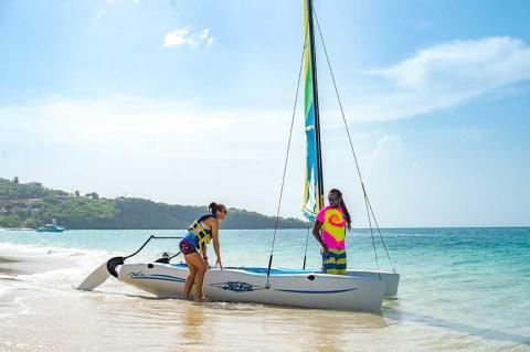Two people set up a colorful sailboat on a tropical beach, ready for adventure.