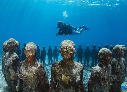 Scuba diver explores submerged statues serving as an artificial reef in clear waters.