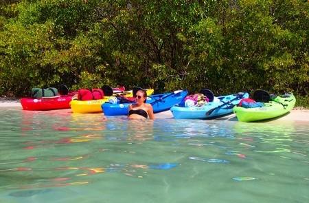Person Relaxing In Clear Water Near Colorful Kayaks Person floating in clear water near beach with colorful kayaks lined up on shore