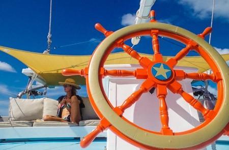 Orange Ship Wheel And Woman On Sunny Boat Deck Orange ships wheel with woman in sun hat on boat under blue sky and yellow sunshade.