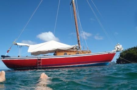Red Sailboat In Tropical Waters With Swimmer Red sailboat with wooden mast in clear blue water, toes visible, sunny tropical setting.