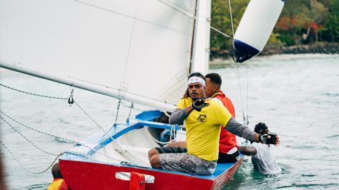 Sailing Competition With Crew On Red And Blue Boat Three men manage sails on a red and blue boat near a green coast.