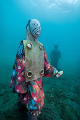 Underwater Tribal Art Scene With Masked Figure Underwater scene with tribal-costumed figure and horned silhouette in background.