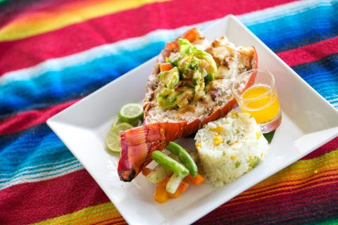 Grilled Lobster Tail With Avocado Salsa And Sides Grilled lobster tail with avocado salsa, rice, vegetables, and dipping sauce on striped fabric.