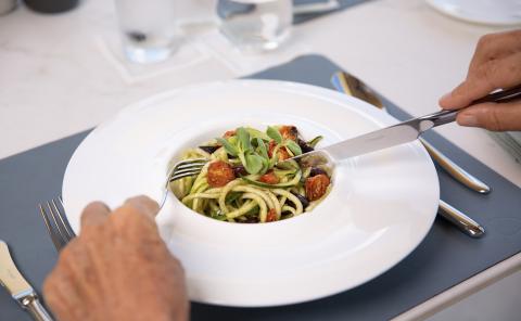 Zucchini Noodles With Sun-Dried Tomatoes And Olives Person eating zucchini noodles garnished with tomatoes and olives on white plate.