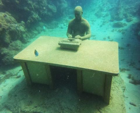 Man Working Underwater With Desk And Fish Man working at desk underwater surrounded by fish and coral, surreal scene.