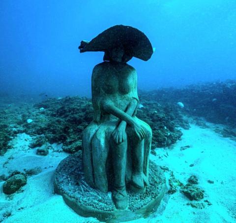 Underwater Statue Art Installation With Marine Life Underwater statue of a seated figure with headdress, surrounded by marine life.