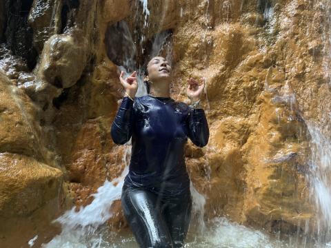 Woman In Wetsuit Enjoying Rocky Waterfall Scene Woman in black wetsuit enjoys serene waterfall with reddish rocks