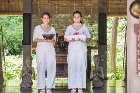 Spa Staff In Traditional Setting With Cultural Decor Two spa staff in white with a wooden bowl and red flower in a serene setting.