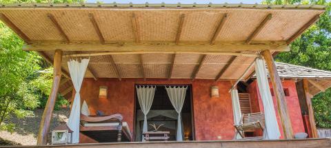 Thatched-roof bungalow with terracotta walls and wooden daybed in lush garden setting