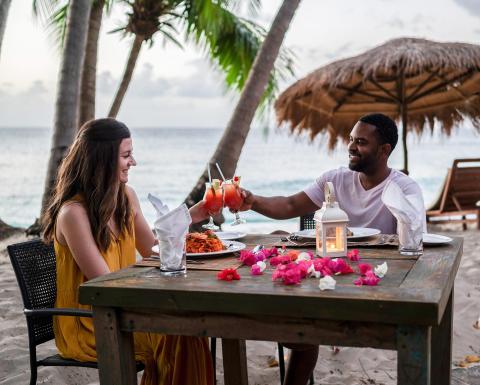 Man and woman toast at beach dinner during sunset with tropical backdrop.