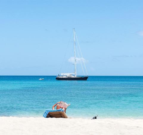 Serene beach with sailboat, motorboat, and person relaxing on sun lounger