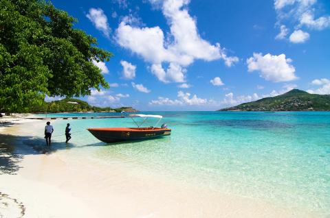 Tropical beach with red boat, clear waters, and people near lush foliage under blue sky.
