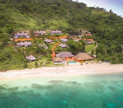 Aerial view of a tropical beachside resort with thatched roofs and lush greenery.