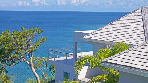 Serene coastal view with modern building, lush tree, and sailboats on blue ocean.