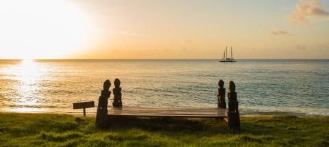 Seaside sunset with couple on bench watching sailboat, peaceful ocean view.