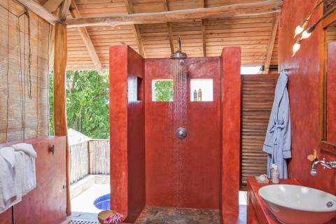 Rustic tropical open-air bathroom with red wall and lush greenery