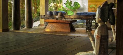 Outdoor patio with wooden daybed, traditional bells, and carved statue, surrounded by plants.