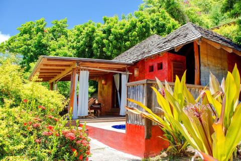 Red cottage with dark roof, bamboo blinds, and lush garden in a tropical setting