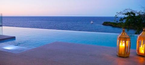 Twilight seascape with infinity pool blending into ocean horizon, sailboat, and lit lanterns.